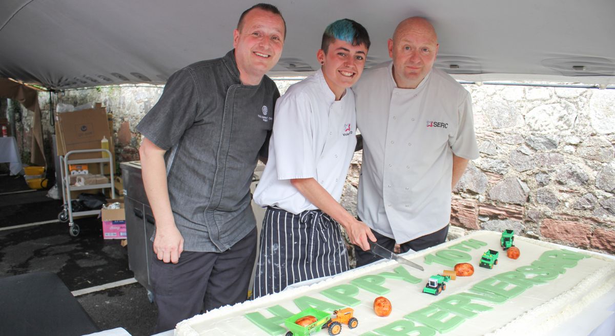 Three chefs stand proudly by large, decorated cake.